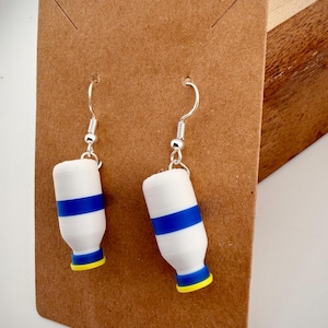 Puede incluir: Un par de pendientes blancos con una franja azul y detalles amarillos, con forma de pequeñas botellas. Cuelgan de ganchos plateados y se exhiben en una tarjeta marrón.