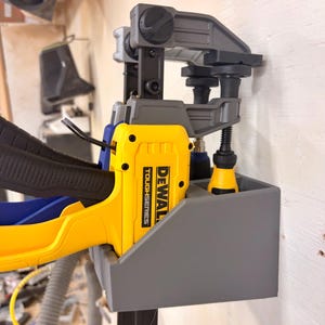 May include: A gray wall-mounted storage bracket holding a yellow and black DeWalt ToughSeries bar clamp. The bracket features a rectangular compartment for the clamp handle and a secondary slot for the clamp head, keeping the tool organized.