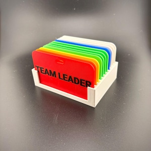 May include: A stack of colorful, rectangular badges in a white holder. The badges are red, orange, yellow, green, blue, and white. The red badge has the words "TEAM LEADER" printed in black.