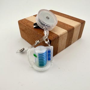 May include: A clear plastic retractable badge reel featuring a coffee maker design. The reel is attached to a wooden box with alternating light and dark brown stripes. The coffee maker design has blue and green accents.