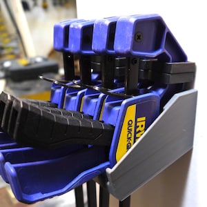 May include: A set of blue and black quick-grip clamps, with the brand name "IRWIN" visible on the side. The clamps are arranged in a holder, with a gray support structure. The background is a workshop setting.