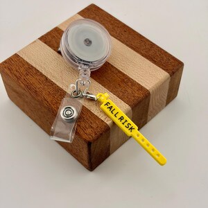 Peut inclure: Un enrouleur de badge r&eacute;tractable en plastique transparent avec un clip m&eacute;tallique et une breloque en silicone jaune en forme de bracelet m&eacute;dical. La breloque porte l'inscription FALL RISK en lettres capitales noires.