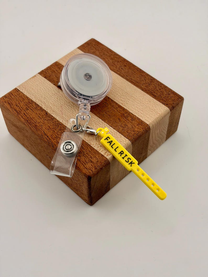 Peut inclure: Un enrouleur de badge r&eacute;tractable en plastique transparent avec un clip m&eacute;tallique et une breloque en silicone jaune en forme de bracelet m&eacute;dical. La breloque porte l'inscription FALL RISK en lettres capitales noires.