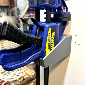 May include: A blue and black Irwin Quick-Grip clamp mounted on a gray holder. The clamp has black handles and a yellow and black label. The holder is attached to a white wall in a workshop setting.
