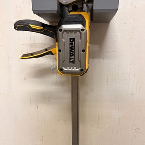 May include: A yellow and black DEWALT clamp with a gray frame. The clamp is mounted on a gray bracket. The DEWALT logo is prominently displayed on the front of the clamp. The clamp has a long, gray bar extending downwards.