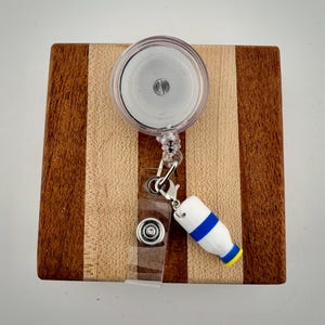 May include: A clear retractable badge reel with a white and blue bottle charm. The reel is attached to a clear plastic strap with a metal snap. The reel is on a wooden block with alternating light and dark wood stripes.