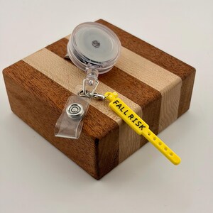 Peut inclure: Un enrouleur de badge r&eacute;tractable dot&eacute; d'un bo&icirc;tier circulaire transparent avec un clip m&eacute;tallique et une sangle en silicone jaune. La sangle jaune porte l'inscription noire FALL RISK et comporte une s&eacute;rie de petits trous circulaires.