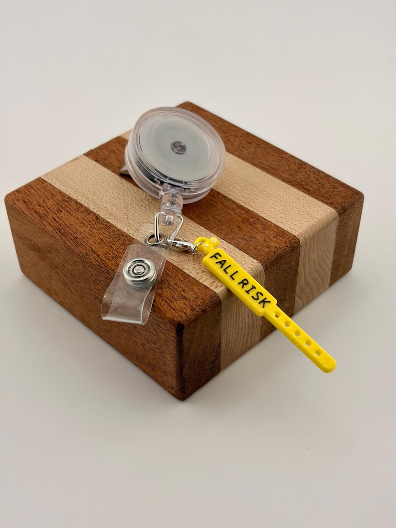 Peut inclure: Un enrouleur de badge r&eacute;tractable dot&eacute; d'un bo&icirc;tier circulaire transparent avec un clip m&eacute;tallique et une sangle en silicone jaune. La sangle jaune porte l'inscription noire FALL RISK et comporte une s&eacute;rie de petits trous circulaires.