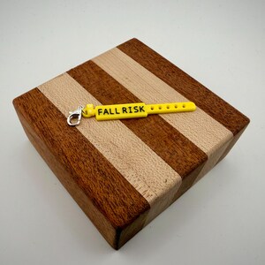 Peut inclure: Une breloque rectangulaire jaune vif en forme de bracelet d'identification m&eacute;dical, avec le texte noir "FALL RISK" imprim&eacute; sur sa surface. La breloque pr&eacute;sente une s&eacute;rie de petites d&eacute;coupes circulaires et un fermoir mousqueton en m&eacute;tal argent&eacute;.