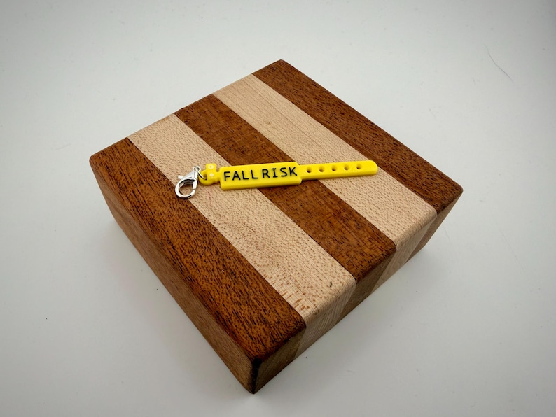 Peut inclure: Une breloque rectangulaire jaune vif en forme de bracelet d'identification m&eacute;dical, avec le texte noir "FALL RISK" imprim&eacute; sur sa surface. La breloque pr&eacute;sente une s&eacute;rie de petites d&eacute;coupes circulaires et un fermoir mousqueton en m&eacute;tal argent&eacute;.