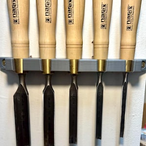 May include: Five chisels with wooden handles and metal blades are displayed in a grey holder. The chisels have a light wood handle, brass ferrule, and dark metal blade. The text "narex Made in Czech Republic" is visible on the handles.