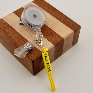 Peut inclure: Un enrouleur de badge r&eacute;tractable en plastique transparent avec un clip m&eacute;tallique et une sangle en vinyle transparent. Attach&eacute; &agrave; l'enrouleur se trouve un pendentif jaune en forme de bracelet m&eacute;dical avec le texte FALL RISK en noir.