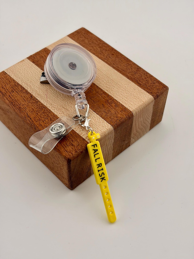 Peut inclure: Un enrouleur de badge r&eacute;tractable en plastique transparent avec un clip m&eacute;tallique et une sangle en vinyle transparent. Attach&eacute; &agrave; l'enrouleur se trouve un pendentif jaune en forme de bracelet m&eacute;dical avec le texte FALL RISK en noir.