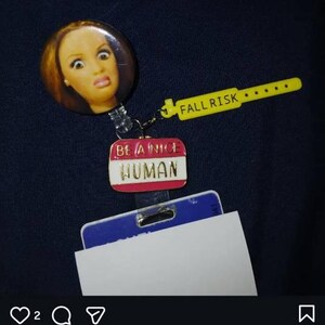 Peut inclure: Un enrouleur de badge avec un bouton circulaire repr&eacute;sentant un visage de poup&eacute;e, une breloque en plastique jaune avec le texte FALL RISK et une breloque &eacute;maill&eacute;e rose et blanche avec le texte BE A NICE HUMAN, fix&eacute; &agrave; un porte-badge bleu et blanc.