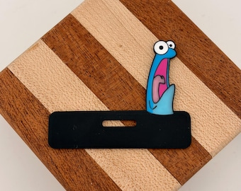 Könnte beinhalten: Ein schwarzer, rechteckiger Ausweishalter aus Silikon mit einer blauen Cartoon-Figur mit großen Augen und offenem Mund. Die Figur ist an der rechten Seite des schwarzen Halters befestigt, der einen horizontalen Schlitz für ein Band besitzt.