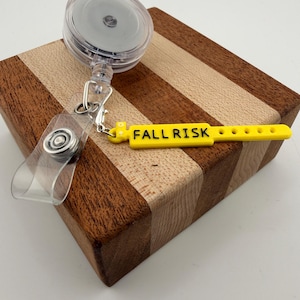 Peut inclure: Un enrouleur de badge r&eacute;tractable en plastique transparent avec un clip m&eacute;tallique et une sangle en silicone jaune. La sangle porte le texte noir FALL RISK imprim&eacute; sur sa surface, ainsi qu'une s&eacute;rie de petits trous circulaires.