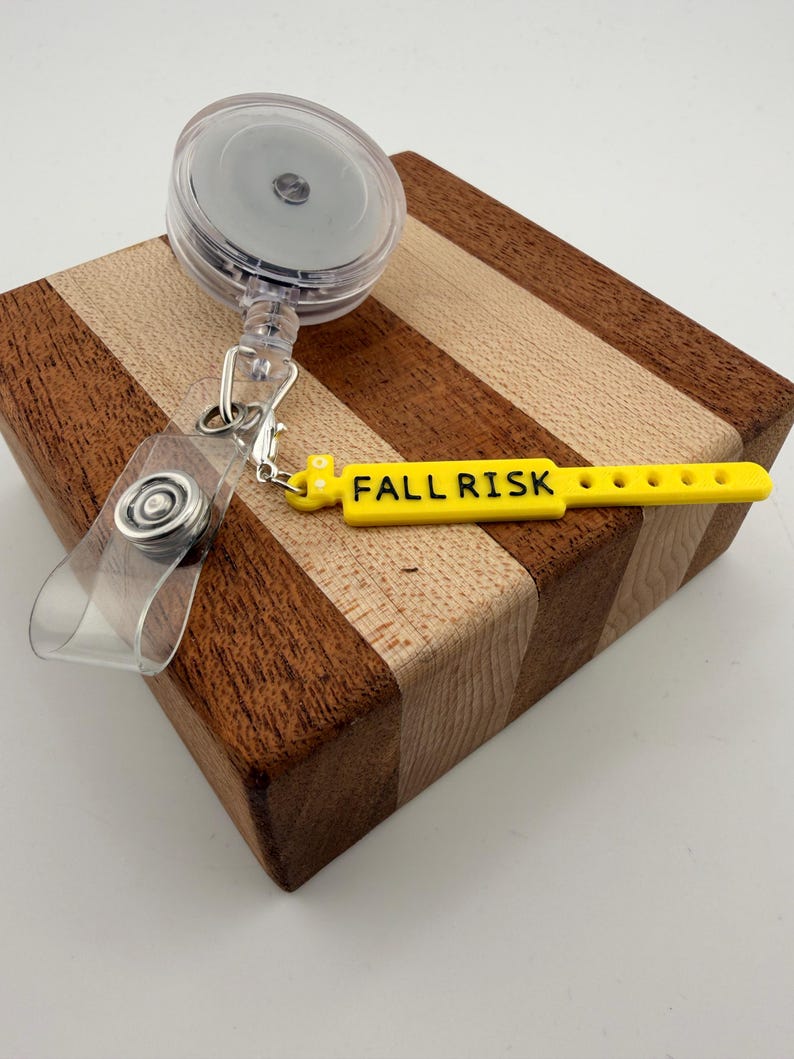 Peut inclure: Un enrouleur de badge r&eacute;tractable en plastique transparent avec un clip m&eacute;tallique et une sangle en silicone jaune. La sangle porte le texte noir FALL RISK imprim&eacute; sur sa surface, ainsi qu'une s&eacute;rie de petits trous circulaires.