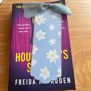 May include: A pale blue bookmark featuring white floral patterns and a grey ribbon, placed on a book titled "House's" by Freida Madden. The book has a purple and blue cover with the text "The New York Times".