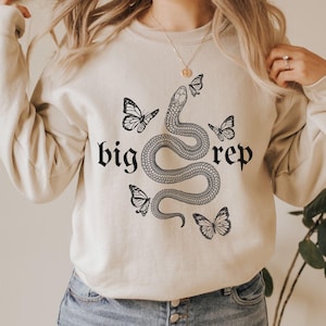 May include: Cream-colored sweatshirt with a black snake and butterfly graphic. The words "big rep" are printed above and below the snake. The sweatshirt has long sleeves and a crew neck.