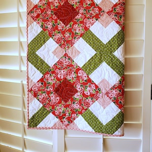 May include: A decorative quilted wall hanging with a geometric pattern. The quilt showcases a central red floral design, framed by white, green, and pink diamond shapes. It has a pink and red patterned border. The quilt is displayed on a white shutter.