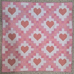 Puede incluir: Un edredón hecho a mano con un patrón repetido de corazones rosas sobre un fondo rosa claro y blanco. El edredón tiene un diseño de patchwork con una mezcla de telas lisas y estampadas, creando un textil decorativo.