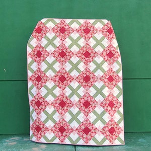 May include: A handmade quilt with a repeating pattern of red floral squares and white and green lattice. The quilt is folded, showcasing the intricate design and color palette. The background is a solid green.