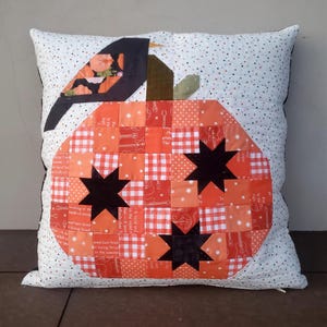 May include: A white pillow with a black bird and a patchwork pumpkin design. The pumpkin is made of orange and red fabric with black stars. The background of the pillow is white with small black dots.