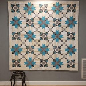 May include: A large square quilt featuring a repeating pattern of bright turquoise eight-pointed stars interspersed with dark gray and white checkered blocks. The background consists of cream-colored fabric strips with a subtle, light-toned pattern.