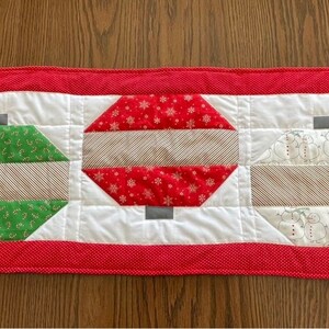 May include: A red, white, and green quilted table runner with a Christmas ornament design. The ornaments are made of different patterned fabrics and have a gray top.