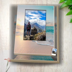 May include: A rectangular picture frame with a light tan border displaying a photograph of a person with a backpack standing on a rocky mountain peak overlooking a beach at sunset. A small miniature version of the same photo is attached to the frame.