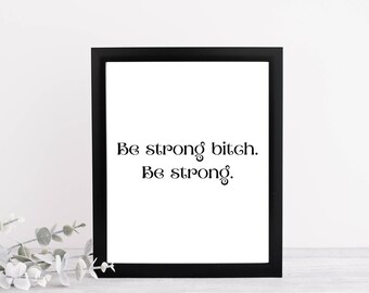 May include: A black framed print displaying the text "Be strong bitch. Be strong." in black script on a white background. The print is on a white surface.