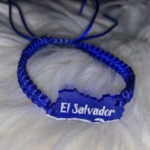 May include: A blue braided bracelet with a blue and white outline of El Salvador. The bracelet is on a white fluffy surface.