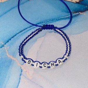 May include: A blue and white friendship bracelet with the word "Bicha" spelled out in white beads.