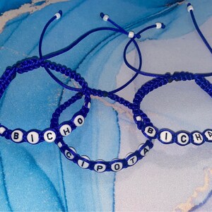 May include: Three blue friendship bracelets with white beads spelling out the words "Bicho", "Cipota", and "Bicha".