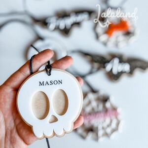 Op de afbeelding: Een wit schedelvormig ornament met de naam "MASON" erop gedrukt. Het ornament heeft een houten achterkant en een zwart touwtje om op te hangen. Andere soortgelijke ornamenten zijn zichtbaar op de achtergrond.