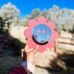 May include: A pink flower-shaped mirror with a smiley face design. The mirror is held in a hand against a blurred background of trees and a blue sky.