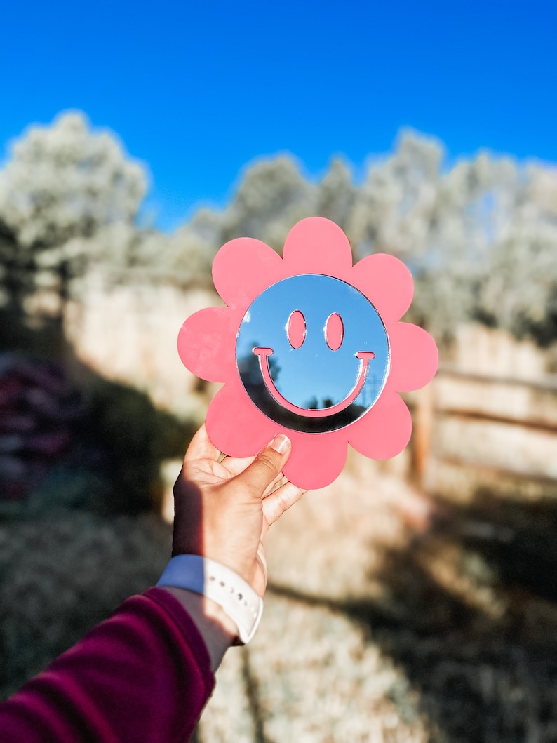 May include: A pink flower-shaped mirror with a smiley face design. The mirror is held in a hand against a blurred background of trees and a blue sky.