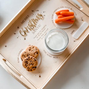 May include: A wooden tray with the words "Merry Christmas Santa!" and "Some Cookies and Some Milk..." engraved on it. The tray also has a small glass jar with milk, a plate with chocolate chip cookies, and a plate with carrots. The tray is decorated with stars and a small white tag that says "Love".