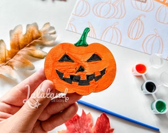 Puede incluir: Una calabaza de madera naranja con un tallo verde y rasgos faciales negros. La imagen incluye botes de pintura, un pincel y una tarjeta que dice "Happy Halloween Pumpkin Patch".