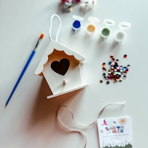 Puede incluir: Una casita de pájaros de madera blanca con una abertura en forma de corazón, botes de pintura, un pincel y lentejuelas de colores. La casita de pájaros tiene una cuerda atada a la parte superior. La etiqueta dice "Muchas gracias".