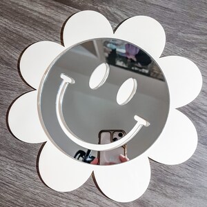 May include: White flower-shaped mirror with a smiley face design. The mirror is reflecting a person holding a phone.