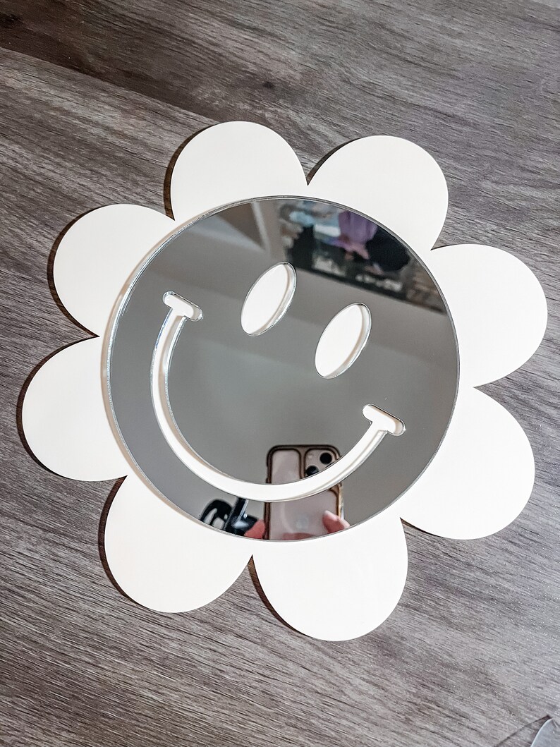 May include: White flower-shaped mirror with a smiley face design. The mirror is reflecting a person holding a phone.
