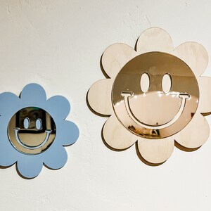 May include: Two wooden flower-shaped mirrors with smiley faces. The larger mirror is natural wood with a gold mirror, and the smaller mirror is light blue with a silver mirror.