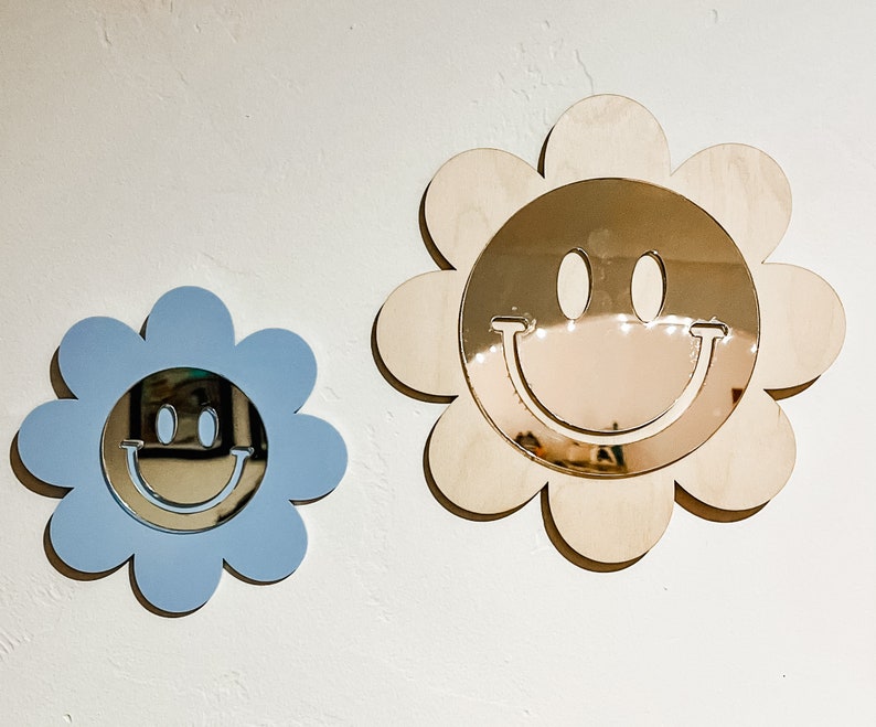 May include: Two wooden flower-shaped mirrors with smiley faces. The larger mirror is natural wood with a gold mirror, and the smaller mirror is light blue with a silver mirror.