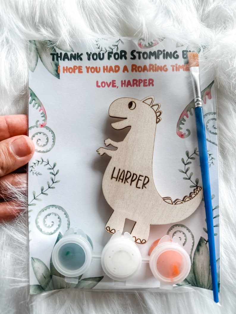 May include: A wooden dinosaur cutout with the name "Harper" painted on it. The dinosaur is surrounded by green leaves and flowers. There are three small paint pots with blue, white, and orange paint. A blue paintbrush is included. The card reads "Thank you for stomping by. I hope you had a roaring time. Love, Harper."