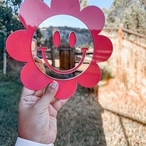 May include: A pink flower-shaped mirror with a smiley face design. The mirror is reflecting a blurry image of a landscape.