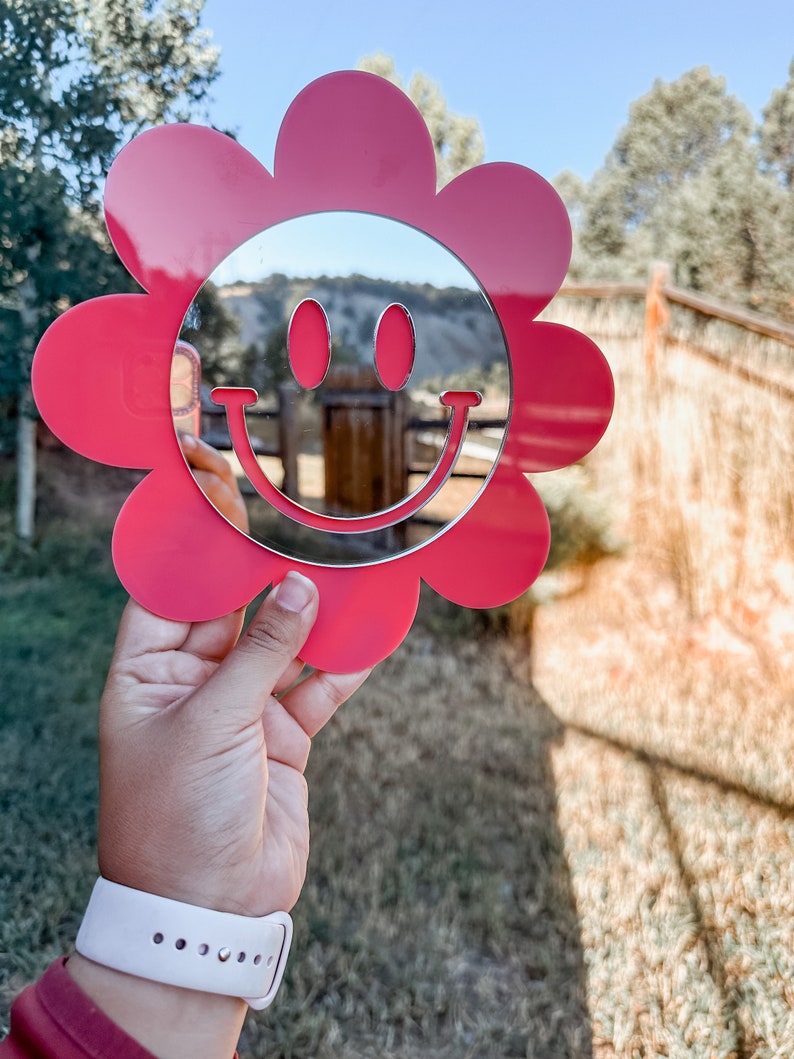 May include: A pink flower-shaped mirror with a smiley face design. The mirror is reflecting a blurry image of a landscape.