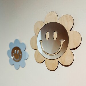 May include: Two wooden flower-shaped wall decor pieces with a mirrored smiley face design. The larger piece is light brown and the smaller piece is light blue.
