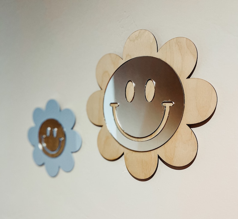 May include: Two wooden flower-shaped wall decor pieces with a mirrored smiley face design. The larger piece is light brown and the smaller piece is light blue.