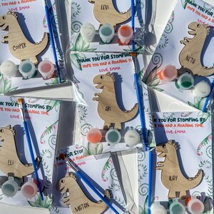 May include: A set of six paint-your-own dinosaur crafts, each with a different name. The dinosaurs are made of wood and have a light brown color. Each craft comes with three small bottles of paint, a paintbrush, and a card with the message "Thank you for stomping by! Hope you had a roaring time. Love, Emma".